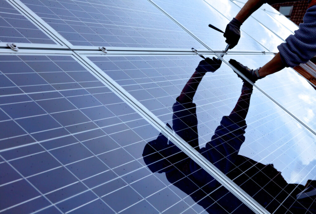 Eine Person montiert blaue Solarmodule, Reflexion sichtbar, Hände werkeln mit Schrauben am Rand.